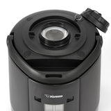A close-up of a black Zojirushi insulated beverage dispenser shows the open lid, revealing a metal mesh filter inside the round opening. The glossy exterior features a visible logo and water level window on the front panel.