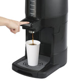 A hand presses a lever on a large insulated beverage dispenser, pouring coffee into a white paper cup placed on the built-in tray. The dispenser features a tall, clear measurement window and a stainless steel vacuum liner to retain heat.