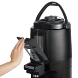 A person’s hand removes a transparent water reservoir from a sleek, black beverage dispenser coffee maker with a curved handle on top. The modern machine features measurement lines on the tank and an insulated, cylindrical design.