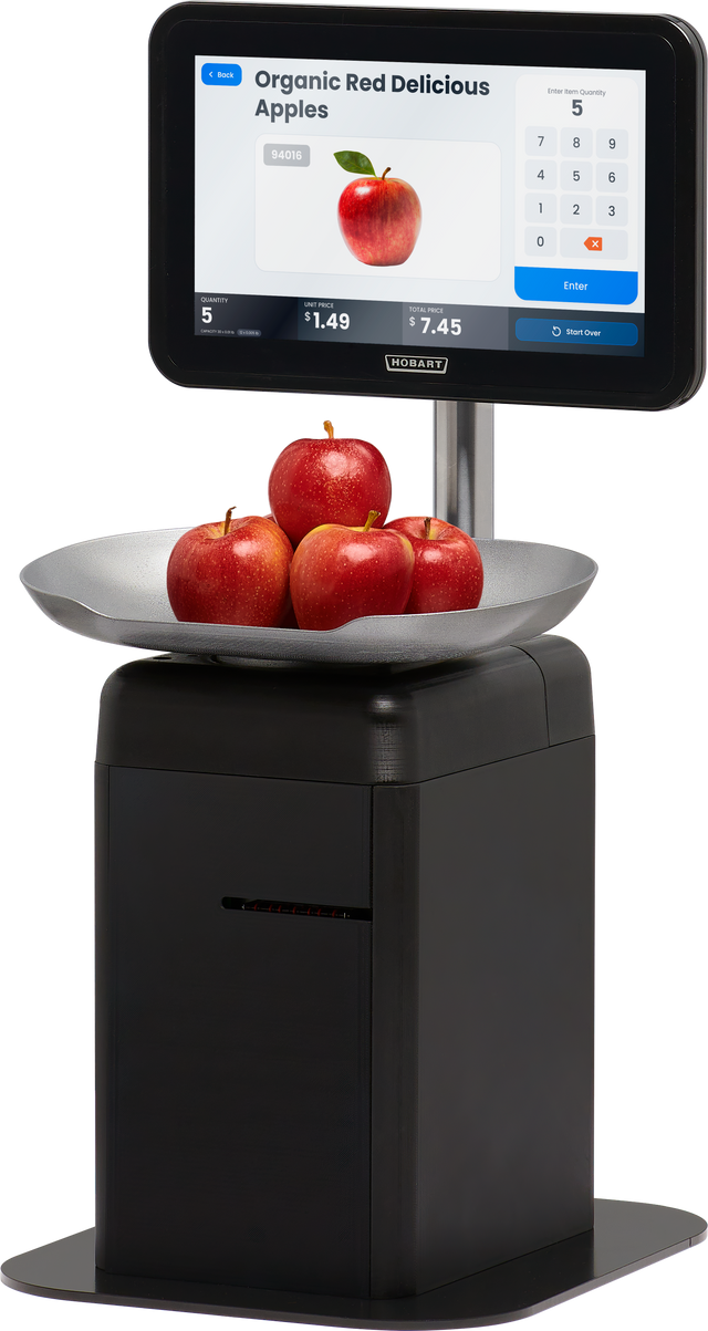 A digital touchscreen scale displays “Organic Red Delicious Apples” with a price and image. Four red apples rest in a metal bowl atop the self-serve scale, which features a black base and a touchscreen for item selection and checkout.