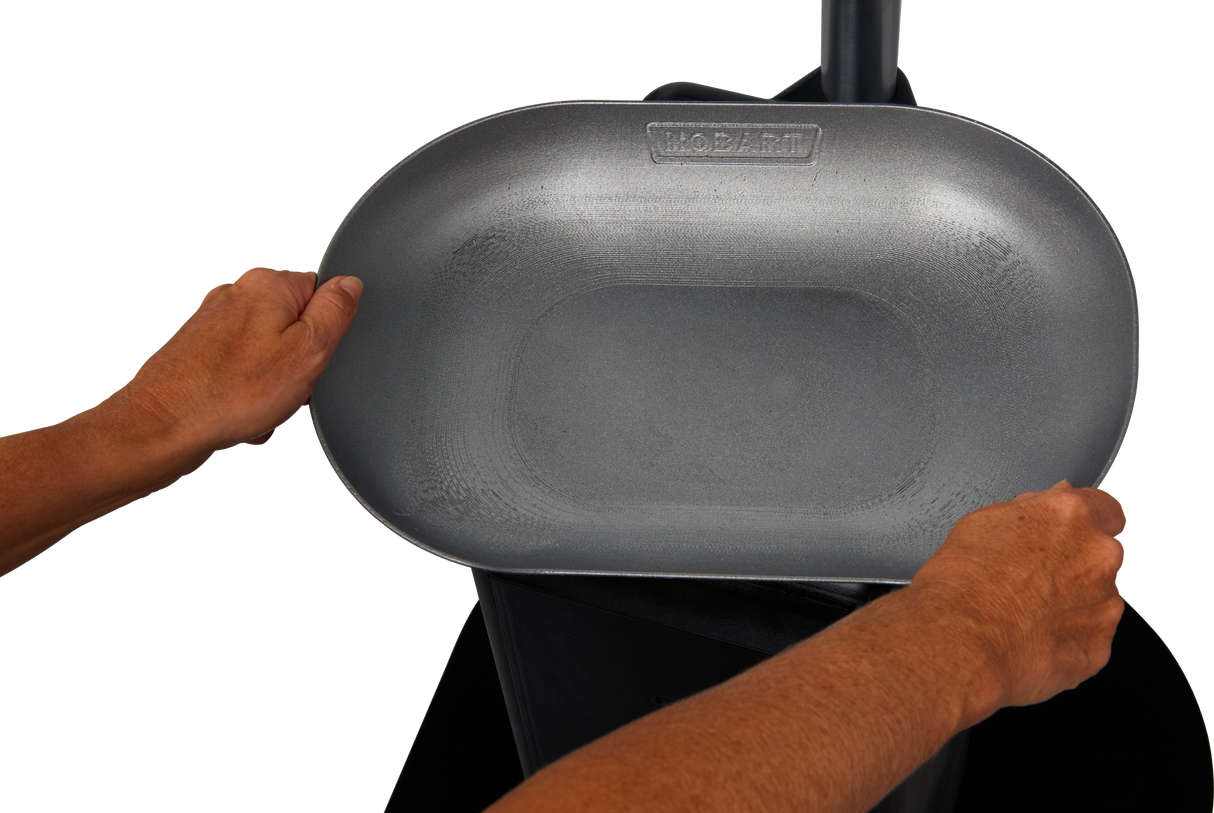 A person’s hands hold a large, gray, oval-shaped metal pan labeled HOBART FS-300 above a black surface, possibly a trash can or appliance, against a plain white background.