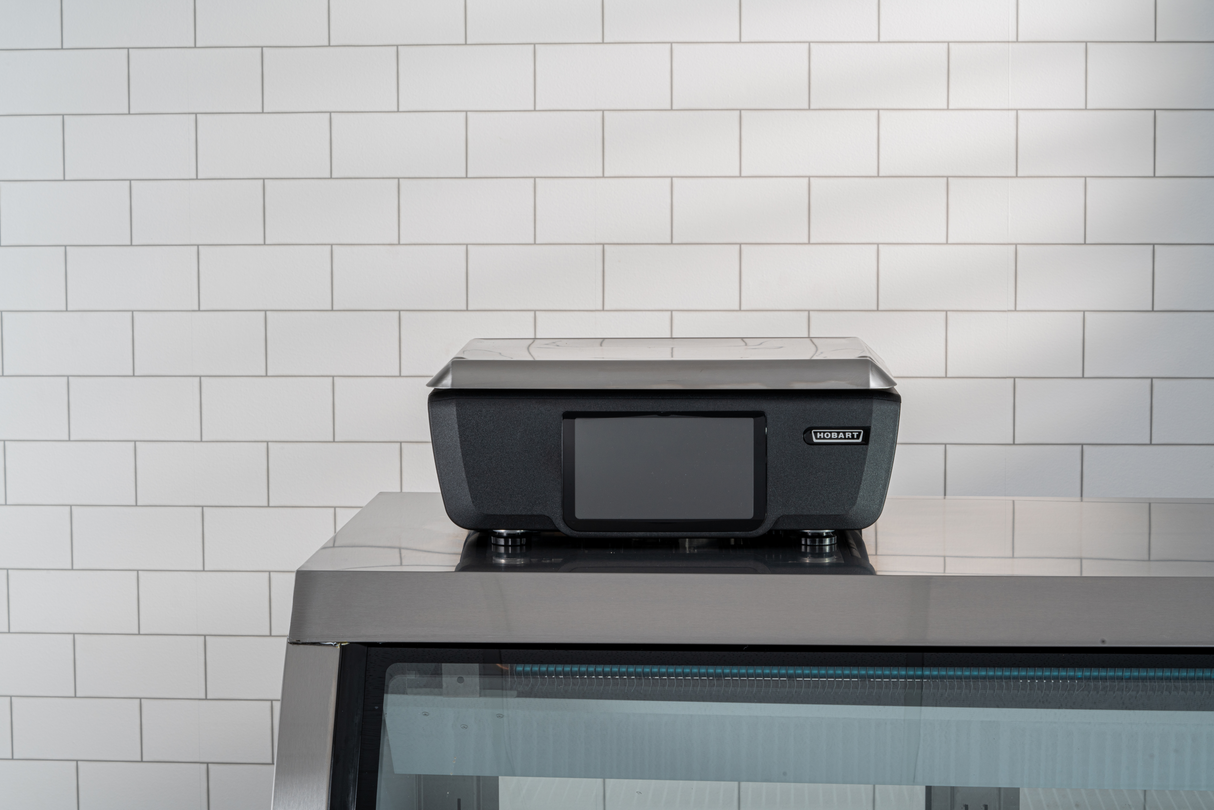 A black GTI Scale/Printer with a closed lid sits atop a glass display case in front of a white tiled wall. The brightly lit scene highlights the printer, while the customer display adds a modern touch to the clean, simple background.