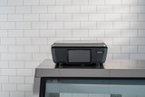 A black GTI Scale/Printer with a closed lid sits atop a glass display case in front of a white tiled wall. The brightly lit scene highlights the printer, while the customer display adds a modern touch to the clean, simple background.