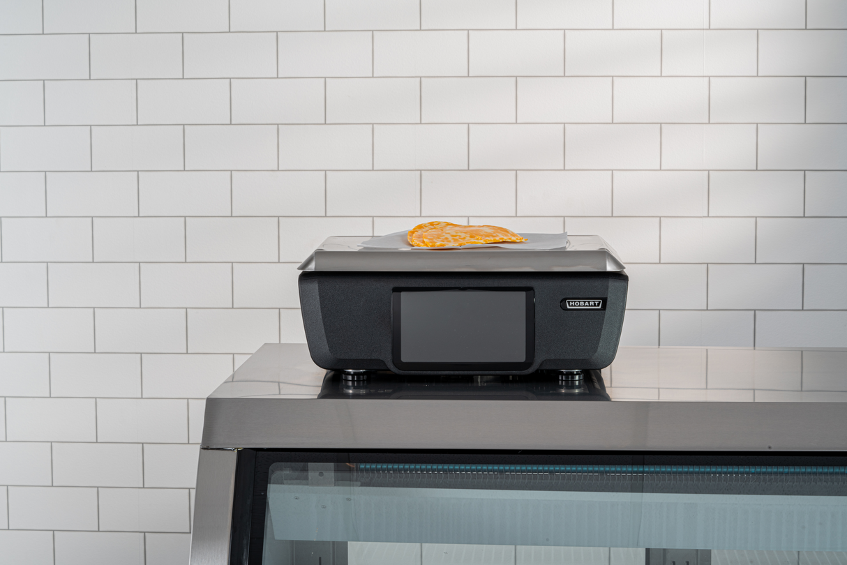 A slice of pizza sits on top of a closed black cash register with a customer display, which is placed on a stainless steel counter. The background features white subway tiles arranged in a grid pattern.