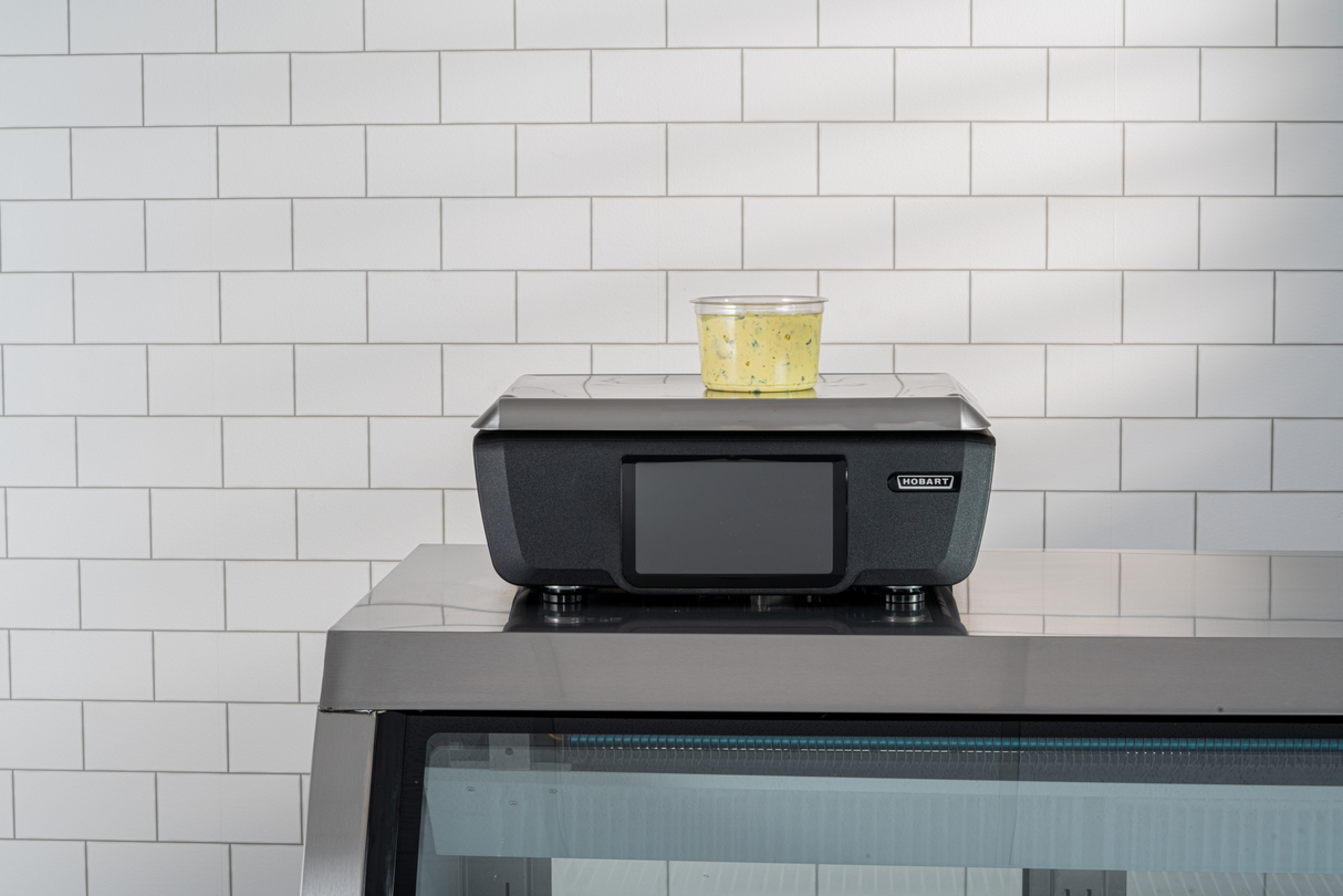 A clear plastic container filled with yellow, herbed food sits atop a black GTI Scale/Printer with a digital screen. The device rests on a stainless steel counter in front of a white subway-tiled wall.
