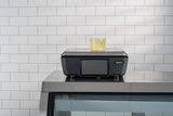 A clear plastic container filled with yellow, herbed food sits atop a black GTI Scale/Printer with a digital screen. The device rests on a stainless steel counter in front of a white subway-tiled wall.