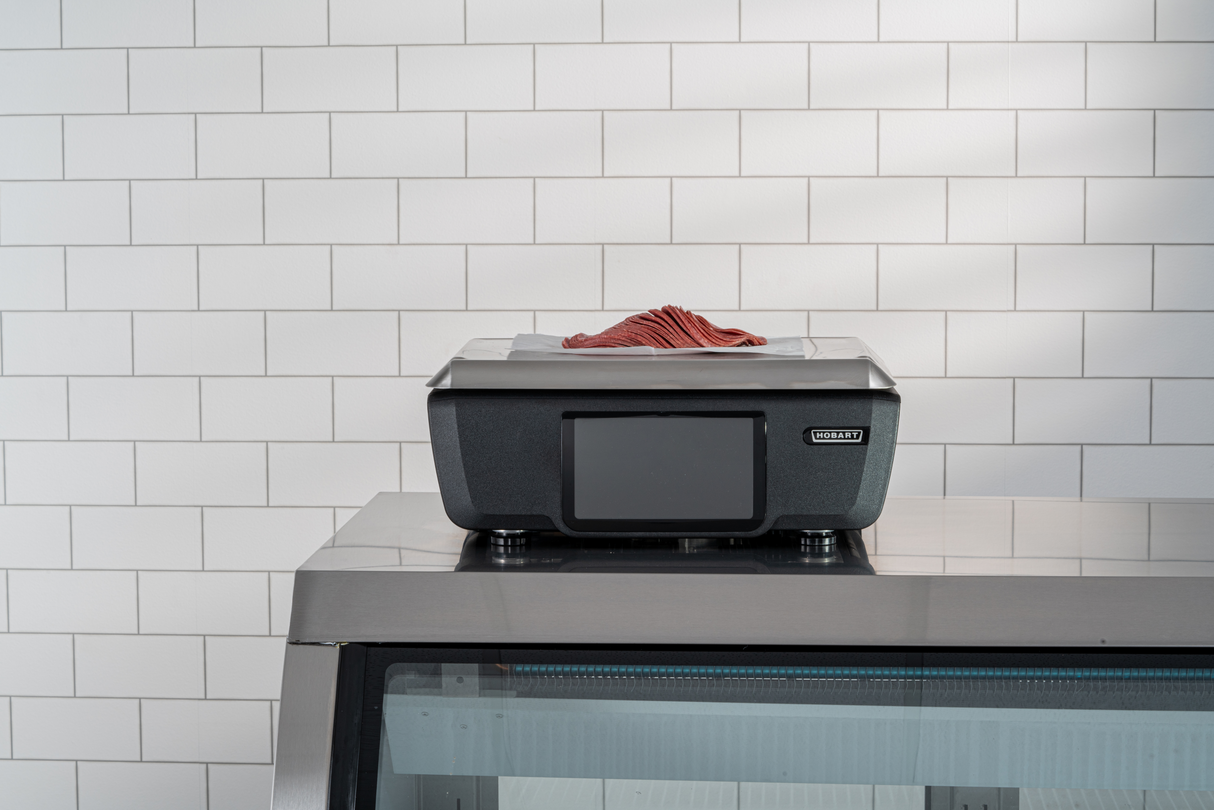 A black Hobart scale with a customer display sits on a stainless steel display case against a white tiled wall. Thin slices of raw red meat are neatly stacked on a tray atop the scale, evoking a clean, minimal butcher or deli setting.