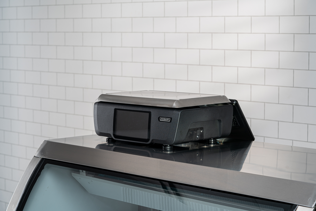 A black and silver rooftop device with a small customer display is mounted on top of a vehicle against a white tiled wall background. The device has a sleek, angular design and appears to be securely attached with visible mounting hardware.