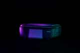 A black GTI Scale/Printer sits on a dark surface against a black background, illuminated by purple and teal lighting that highlights its contours. The printer has a flatbed top and rectangular front panel, giving it a modern, sleek appearance.