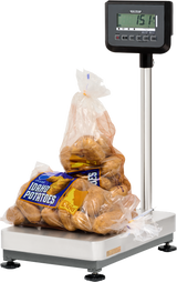 A plastic bag of Idaho potatoes sits on a legal for trade bench receiving scale with a 300 lb capacity. The display reads 15.14 pounds, and the partially open bag reveals several brown potatoes against a plain white background.