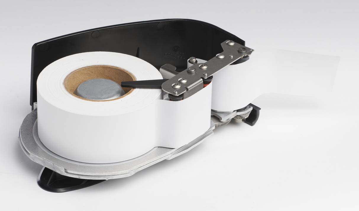 A close-up of a label dispenser with a white roll of labels loaded inside. The device features a black and silver body with an auto cutter mechanism feeding and slicing the label strip. Unused linerless cassette labels are seen coming out on the right side.