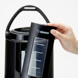 A hand removes a black, rectangular Zojirushi water tank with a clear window and level markings from an insulated beverage dispenser featuring a curved handle and cylindrical stainless steel body, shown against a plain white background.