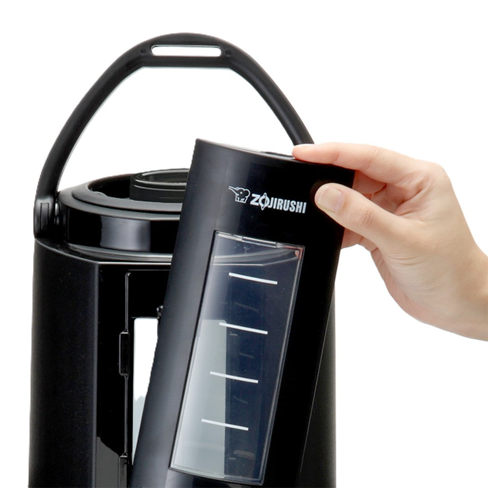 A hand removes a black, rectangular water-level gauge from an insulated Zojirushi beverage dispenser. The gauge has a transparent window with white markings, and the main appliance features a handle and open top for easy access.