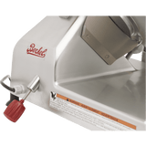 Close-up of a silver Berkel slicer, a manual gravity feed slicer, showing the brand logo, red-handled control knob, metal lever, and an orange warning label with text in English, French, and Spanish on the machine’s shiny metal surface.