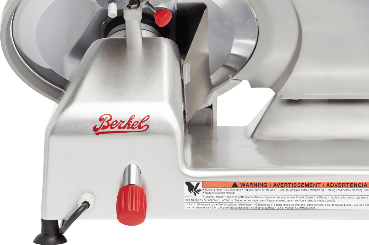 Close-up of a manual gravity feed slicer with the brand name Berkel in red script. The image shows a red knob, blade cover, safety guard, and warning label in three languages. The slicer appears clean and unused.