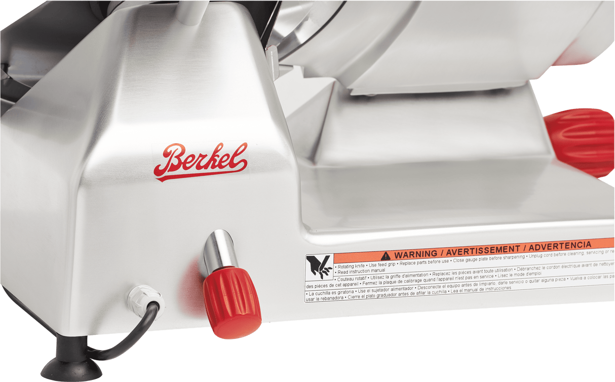 Close-up of a stainless steel Berkel manual gravity feed slicer. The red Berkel logo and two red knobs are visible, along with a carbon steel knife blade, black power cord at the base, and a warning label.