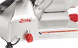 Close-up of a stainless steel Berkel manual gravity feed slicer. The red Berkel logo and two red knobs are visible, along with a carbon steel knife blade, black power cord at the base, and a warning label.