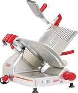 A manual gravity feed slicer with a tilted stainless steel platform, carbon steel knife, large circular blade, red handles, and adjustment knobs. Designed for slicing deli meats and cheeses, it features safety guards and front control switches.