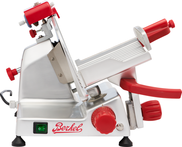 A stainless steel Berkel gravity feed slicer with red knobs and handles, a tilted adjustable platform, and a sharp circular blade. The machine has an on/off switch and the Berkel logo in red. Its overall design is modern and industrial.