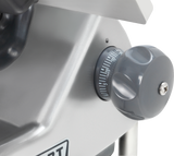 A close-up of a gray rotary dial on a heavy duty meat slicer, marked with numbers from 0 to 15 in white. The ridged dial is mounted on a smooth, angled metallic surface with visible screws, while the background remains out of focus.