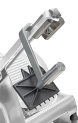 Close-up of a commercial Hobart meat slicer, featuring a heavy duty meat grip with metal spikes on a black plastic holder. The sharp circular blade, safety guard, and textured sliding platform highlight its clean, metallic design.