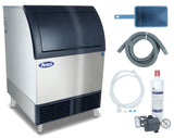 A silver cube-style ice machine with a black lid sits left. To the right, a blue scoop, gray hose, white tube, water filter, and small gauge are arranged. The Ice Maker features the Atosa logo on its stainless steel ice bin front.