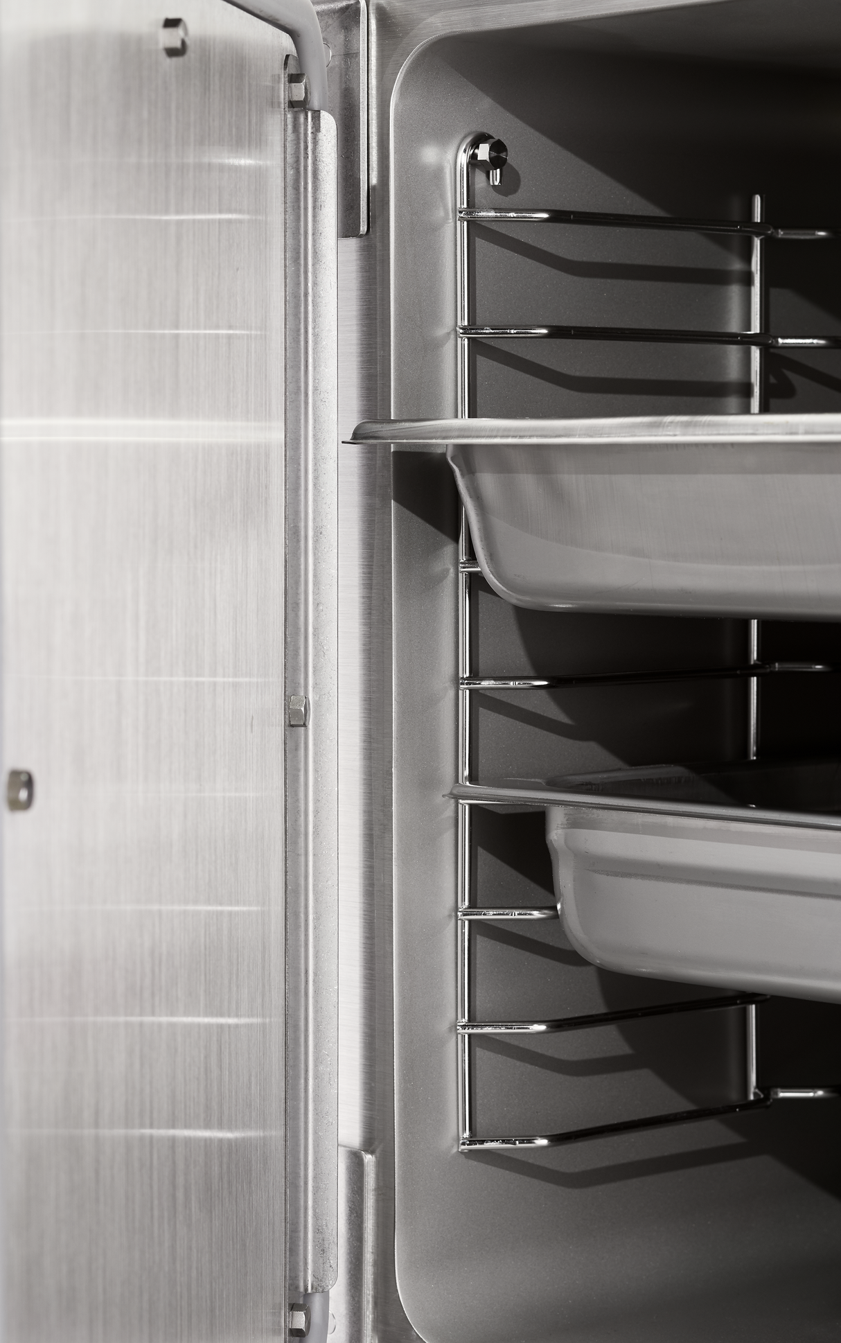 Close-up of a stainless steel convection steamer interior, featuring metal racks and two shallow trays on different levels. The left side displays the brushed metal door with a visible hinge. The scene is well-lit and clean.