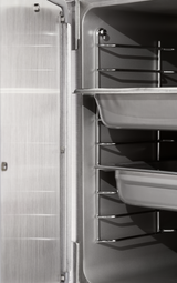 Close-up of a stainless steel convection steamer interior, featuring metal racks and two shallow trays on different levels. The left side displays the brushed metal door with a visible hinge. The scene is well-lit and clean.
