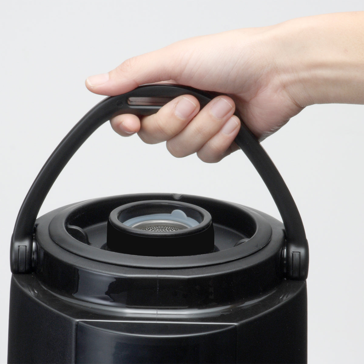 A close-up of a hand gripping the black plastic handle of a large, round insulated beverage dispenser. The container has a circular lid with a central spout or opening, and the background is plain and light-colored.