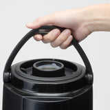 A close-up of a hand gripping the black plastic handle of a large, round insulated beverage dispenser. The container has a circular lid with a central spout or opening, and the background is plain and light-colored.