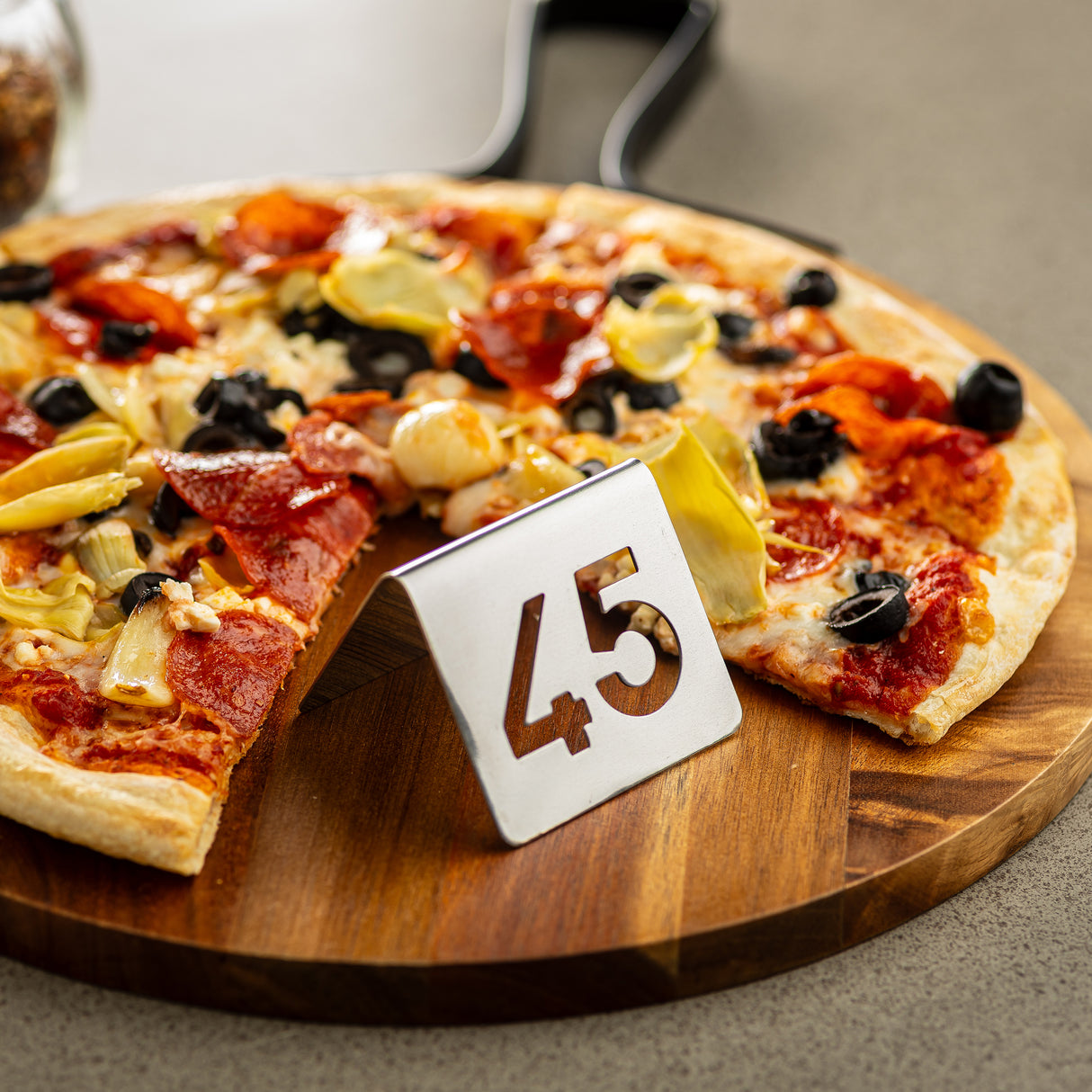A pizza topped with pepperoni, artichokes, black olives, and cheese sits on a wooden board. In front of the pizza is a TableCraft stainless steel number tent set with the number 45 cut out. The background is blurred.