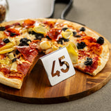 A pizza topped with pepperoni, artichokes, black olives, and cheese sits on a wooden board. In front of the pizza is a TableCraft stainless steel number tent set with the number 45 cut out. The background is blurred.