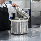 A person in a white chef coat and black apron lifts the lid of a large Zojirushi America commercial rice cooker on a reflective kitchen counter, set amid metal appliances and storage units in a bustling commercial kitchen.