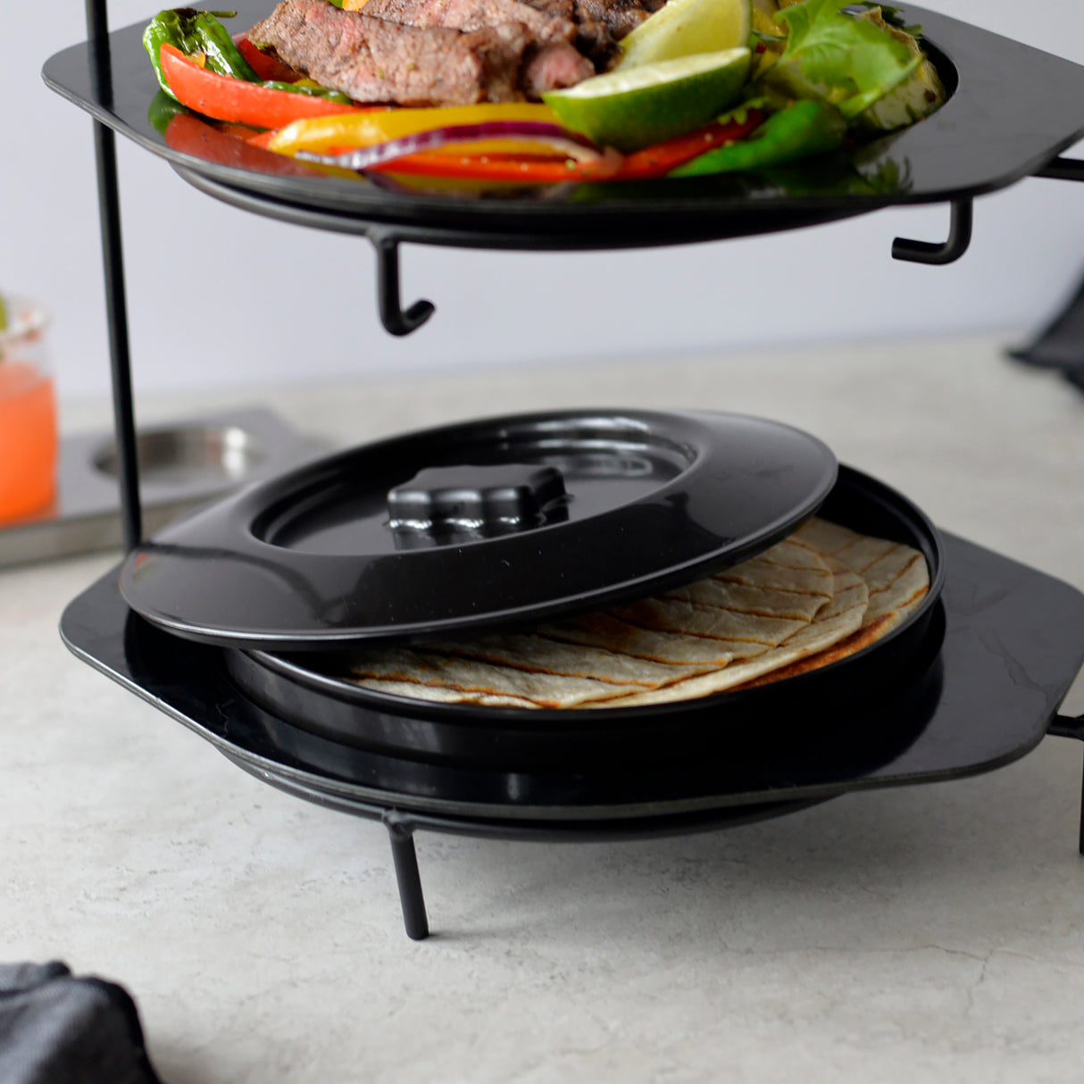 A two-tiered black metal serving stand holds grilled meat and vegetables on the top plate. Below, a lidded polypropylene Tortilla Warmer keeps tortillas fresh. The setup is on a light gray countertop, with a drink and napkin in the background.