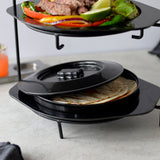 A two-tiered black metal serving stand holds grilled meat and vegetables on the top plate. Below, a lidded polypropylene Tortilla Warmer keeps tortillas fresh. The setup is on a light gray countertop, with a drink and napkin in the background.