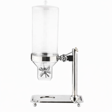 The TableCraft Cereal Dispenser features a clear cylindrical container, sleek stainless steel design, and a convenient paddle handle for easy dispensing. Its sturdy rectangular base with circular cutout and round feet adds a modern touch.