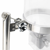 Close-up of a TableCraft cereal dispenser with a shiny black adjustment knob and bracket securing it to a stainless steel pole. The clear cylindrical container above is paired with a paddle handle for easy dispensing.