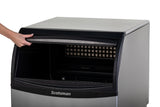 A person’s hand lifts the black front door of a stainless steel Scotsman undercounter ice maker, revealing the interior ice bin and ice outlet grid. The plain white background highlights this industrial appliance.