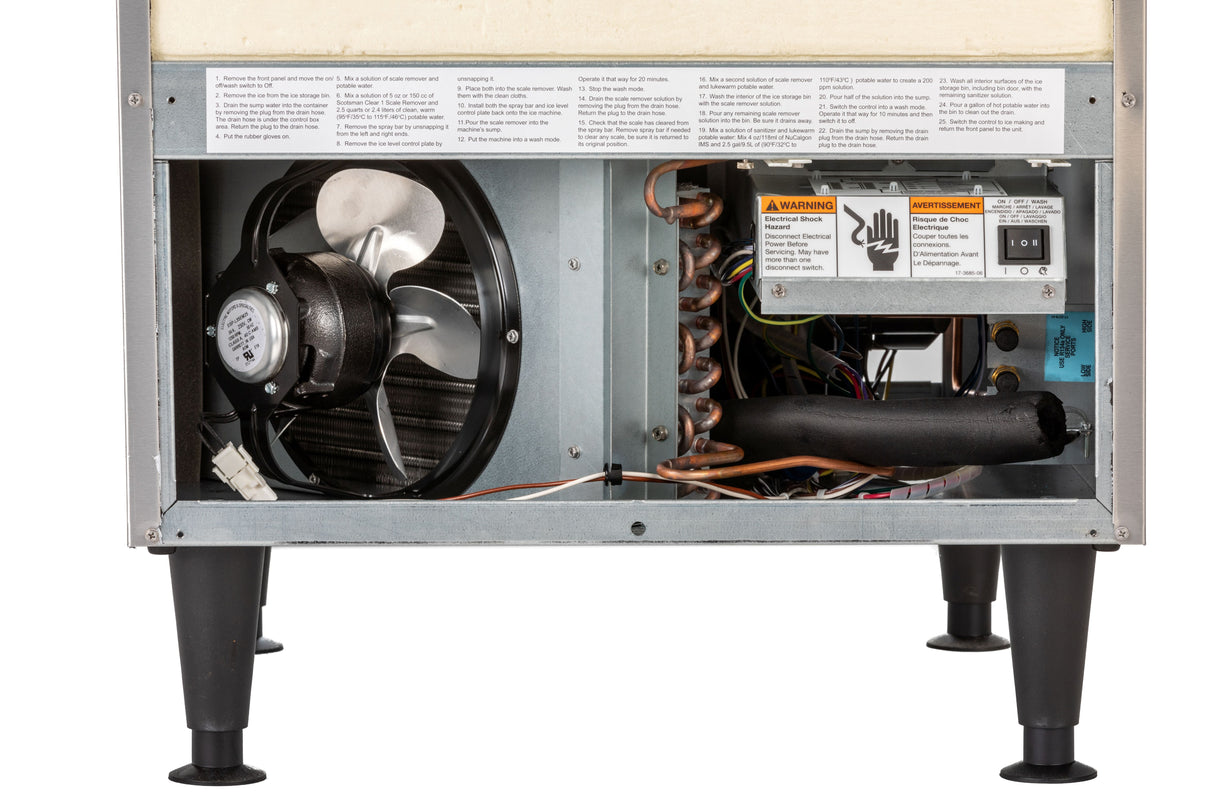 Close-up of the lower section of an undercounter ice maker with its service panel removed, revealing a fan, copper coils, wires, warning labels, a control box, and black insulation tubing—all supported by four black legs.