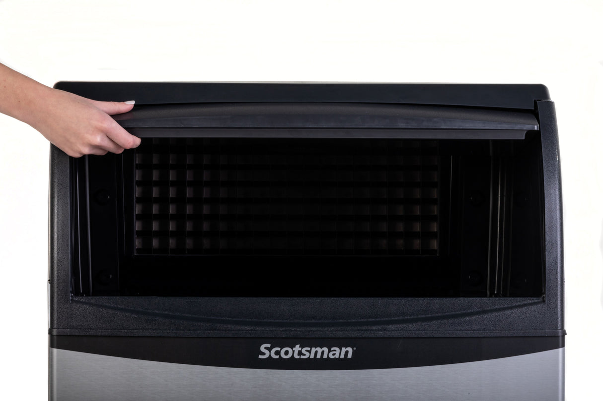A hand lifts the top lid of a Scotsman undercounter ice maker, revealing the empty black interior and spacious ice bin storage. The stainless steel front displays the Scotsman brand at the bottom against a plain white background.