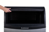 A hand lifts the top lid of a Scotsman undercounter ice maker, revealing the empty black interior and spacious ice bin storage. The stainless steel front displays the Scotsman brand at the bottom against a plain white background.