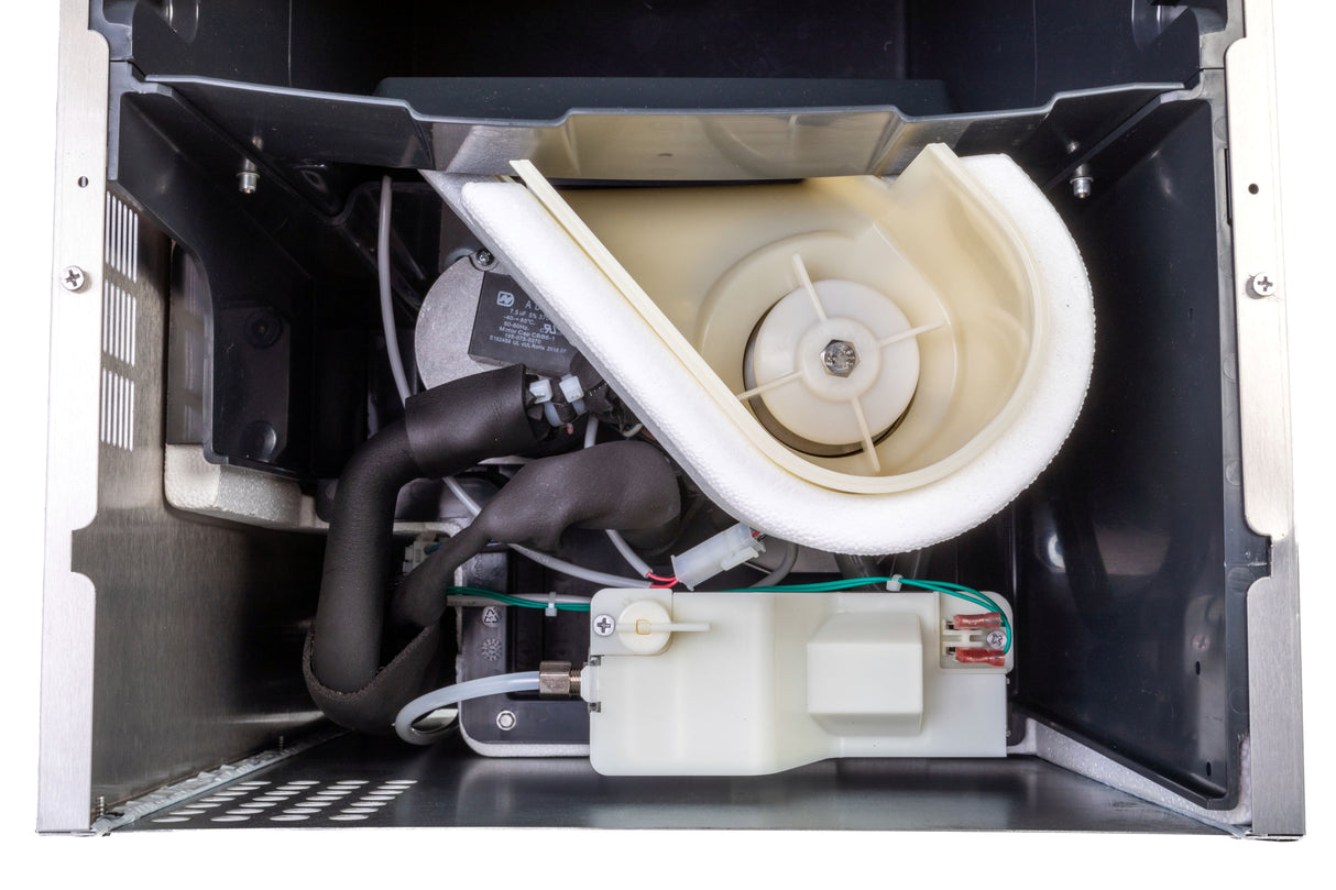 Inside view of an ADA Compliant undercounter ice maker showing internal components: a white plastic water reservoir with wires, black hoses, a fan-like impeller, and various metal and plastic parts inside a metal housing, illustrating the water and cooling system.