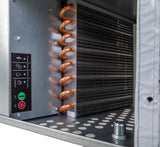 Close-up of an industrial HVAC evaporator coil with copper tubing and aluminum fins, resembling components in an undercounter ice maker. A control panel with status indicators sits on the left, while the perforated metal base features a protruding bolt.