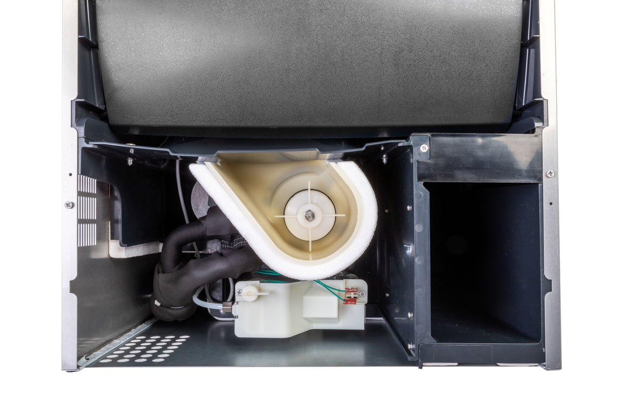 Close-up view of the inside of an undercounter ice maker, showing various internal components including a white plastic blower housing, insulated pipes, wiring, and a black metal compartment—all housed within a metallic structure.