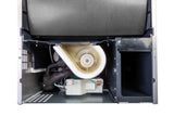 Close-up view of the inside of an undercounter ice maker, showing various internal components including a white plastic blower housing, insulated pipes, wiring, and a black metal compartment—all housed within a metallic structure.