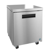 A Hoshizaki undercounter refrigerator with a single solid front door, black handle, and compact square shape sits on four caster wheels. Its smooth stainless steel surface gleams, accented by a small blue certification sticker in the lower right corner.