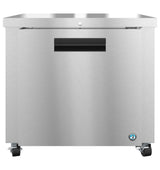A stainless steel undercounter refrigerator or freezer unit with a flat top, black handle, small lock above the handle, and four caster wheels. The brushed metal finish is complemented by a round blue manufacturer logo on the bottom right corner.