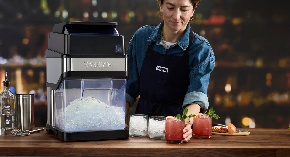 A person in a denim shirt and apron stands at a bar, using a commercial ice crusher filled with crushed ice. Beside it are glasses with ice and two pink cocktails garnished with mint, set on a wooden counter with cocktail tools and fruit.