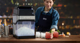 A person in a denim shirt and apron stands at a bar, using a commercial ice crusher filled with crushed ice. Beside it are glasses with ice and two pink cocktails garnished with mint, set on a wooden counter with cocktail tools and fruit.