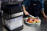 A person in a Waring apron prepares a platter of assorted sashimi on crushed ice beside a Waring commercial ice crusher on a stainless steel countertop in a commercial kitchen setting.